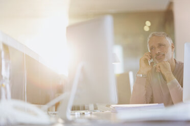 Businessman talking on cell phone at computer in sunny office - HOXF00376