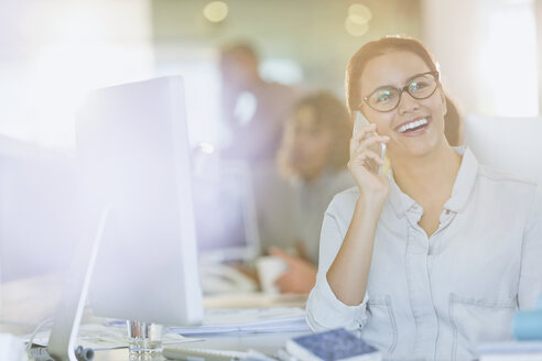 Lächelnde Geschäftsfrau im Gespräch mit Handy am Computer im Büro - HOXF00412