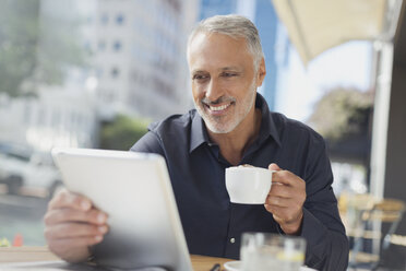 Lächelnder Geschäftsmann mit digitalem Tablet, der in einem Straßencafé Kaffee trinkt - HOXF00524