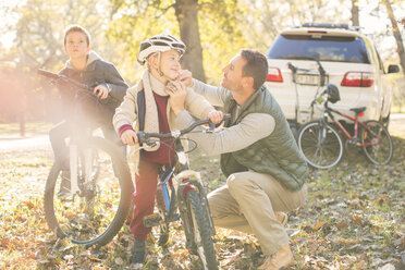 Father fastening helmet of son on bicycle in autumn woods - HOXF00619