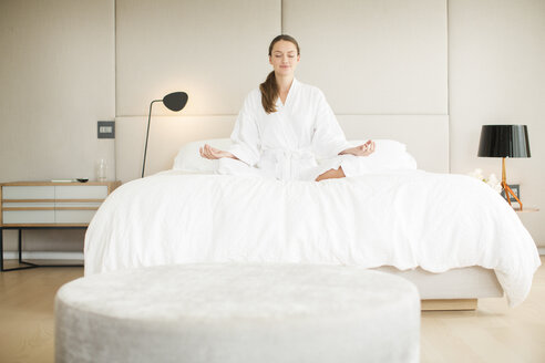 Gelassene Frau im Bademantel meditiert im Lotussitz auf dem Bett - HOXF00756