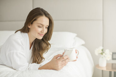 Lächelnde Frau im Bademantel, die auf dem Bett Kaffee trinkt und eine SMS auf ihrem Handy schreibt - HOXF00757