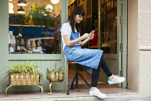 Woman sitting on stool using cell phone at entrance door of a store - EBSF02240