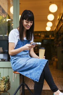 Lächelnde Frau, die auf einem Hocker sitzt und ein Mobiltelefon an der Eingangstür eines Geschäfts benutzt - EBSF02241