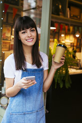 Portrait of smiling woman with cell phone and takeaway coffee in entrance door of a store - EBSF02242