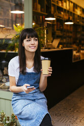 Smiling woman sitting at entrance door of a store holding cell phone and takeaway coffee - EBSF02243