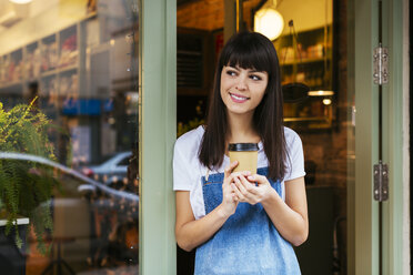 Smiling woman standing at entrance door of a store holding takeaway coffee - EBSF02245
