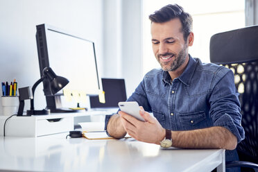 Lächelnder Mann benutzt Handy am Schreibtisch im Büro - BSZF00240