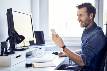 Smiling man using cell phone at desk in office - BSZF00243