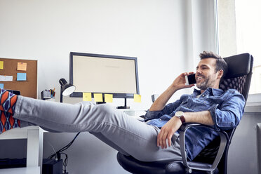 Entspannter Mann sitzt am Schreibtisch im Büro und spricht mit seinem Handy - BSZF00254