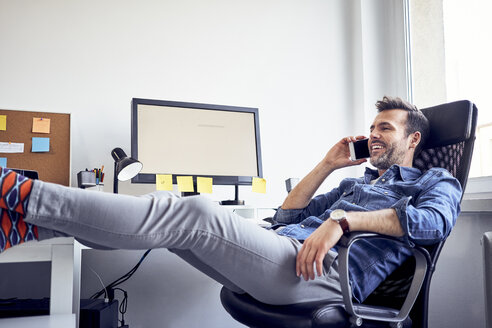 Entspannter Mann sitzt am Schreibtisch im Büro und spricht mit seinem Handy - BSZF00254