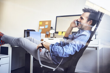 Entspannter Mann sitzt am Schreibtisch im Büro und spricht mit seinem Handy - BSZF00255