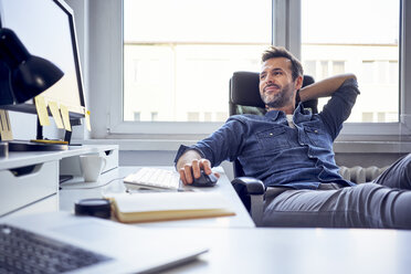 Relaxed man sitting at desk in office looking at computer screen - BSZF00256