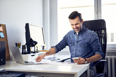 Smiling man with smartphone and draft working at desk in office - BSZF00264