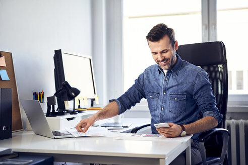Lächelnder Mann mit Smartphone und Entwurf arbeitet am Schreibtisch im Büro - BSZF00264