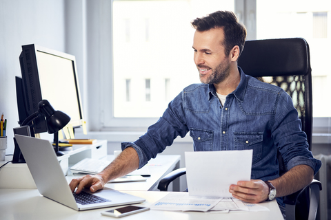 Lächelnder Mann hält ein Dokument und benutzt einen Laptop am Schreibtisch im Büro, lizenzfreies Stockfoto