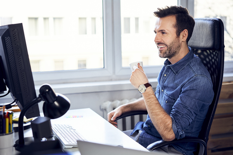 Lächelnder Mann schaut auf den Computer und trinkt Kaffee am Schreibtisch im Büro, lizenzfreies Stockfoto