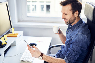 Smiling man looking at cell phone and drinking coffee at desk in office - BSZF00269