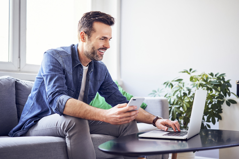 Lächelnder Mann auf dem Sofa sitzend mit Laptop und Mobiltelefon, lizenzfreies Stockfoto