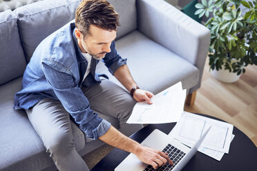 Man with documents sitting on sofa using laptop - BSZF00281