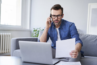 Serious man with documents and laptop on sofa talking on cell phone - BSZF00284