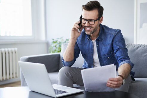 Smiling man with documents and laptop on sofa talking on cell phone - BSZF00285