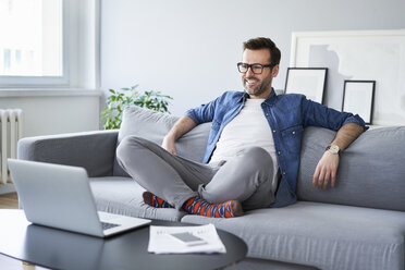 Relaxed smiling man sitting on sofa looking at laptop - BSZF00286