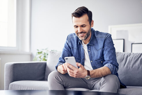 Lächelnder Mann sitzt auf dem Sofa und benutzt ein Mobiltelefon - BSZF00291