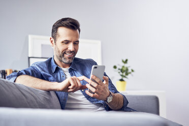 Smiling man sitting on sofa using cell phone - BSZF00292