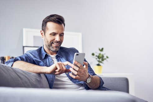 Lächelnder Mann sitzt auf dem Sofa und benutzt ein Mobiltelefon - BSZF00292
