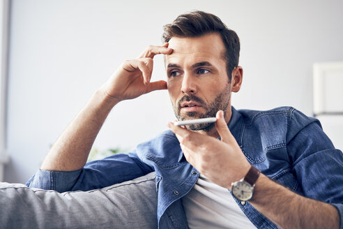 Worried man sitting on sofa using cell phone - BSZF00294