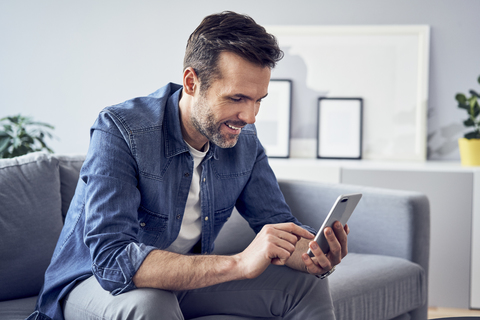 Lächelnder Mann sitzt auf dem Sofa und benutzt ein Mobiltelefon, lizenzfreies Stockfoto