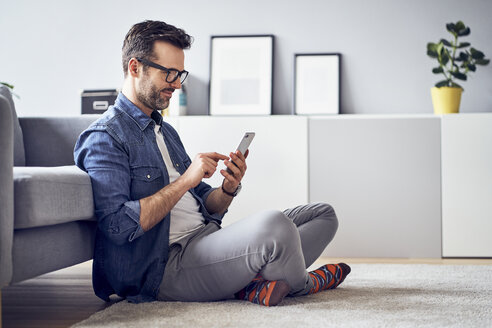 Lächelnder Mann, der im Wohnzimmer auf dem Boden sitzt und ein Mobiltelefon benutzt - BSZF00300