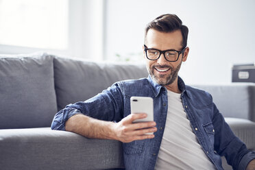 Lächelnder Mann im Wohnzimmer, der auf sein Handy schaut - BSZF00301