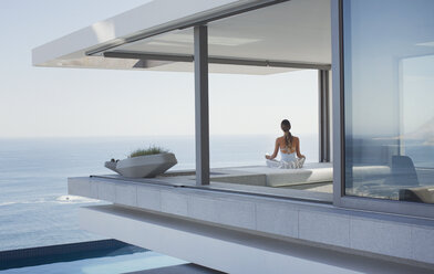 Gelassene Frau meditiert auf einer modernen, luxuriösen Musterhaus-Terrasse mit Meerblick - HOXF01027