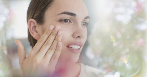 Brunette woman applying face moisturizer to cheek - HOXF01036