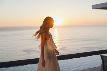 Frau im weißen Kleid auf einer ruhigen, luxuriösen Terrasse mit Blick auf das Meer bei Sonnenuntergang - HOXF01040