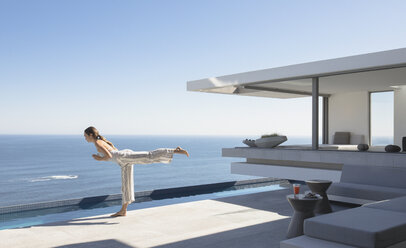 Frau übt Yoga Krieger 3 Pose auf sonnigen modernen, luxuriösen Haus Schaufenster Außenterrasse mit Meerblick - HOXF01042