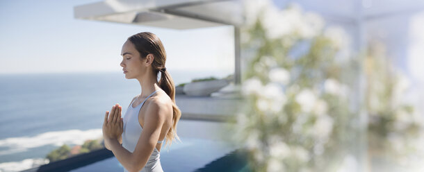 Gelassene brünette Frau, die Yoga praktiziert und mit den Händen in der Herzmitte auf einer sonnigen Terrasse mit Blick auf das Meer meditiert - HOXF01059