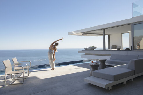 Frau übt Yoga Seite dehnen auf moderne, luxuriöse Haus Schaufenster Außenterrasse mit sonnigen Blick auf das Meer - HOXF01066