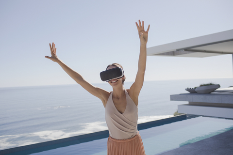 Energetische Frau, die eine Virtual-Reality-Simulator-Brille auf einer modernen, luxuriösen Wohnung mit Blick auf das Meer benutzt, lizenzfreies Stockfoto