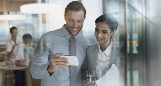 Lächelnder Geschäftsmann und Geschäftsfrau, die ein Video auf einem Smartphone im Büro ansehen - HOXF01152
