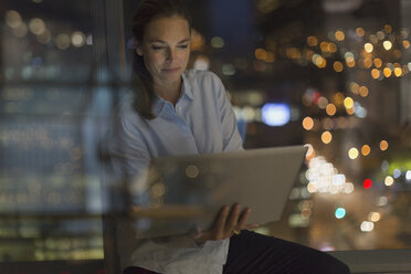 Serious businesswoman working late at laptop in office window at night - HOXF01155