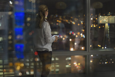 Nachdenkliche Geschäftsfrau mit digitalem Tablet, die spät arbeitet und nachts aus dem Fenster eines städtischen Büros schaut - HOXF01163