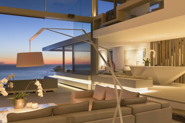 Illuminated, modern luxury home showcase interior living room with ocean view at dusk - HOXF01248