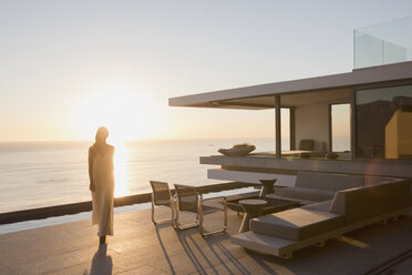 Frau, die auf der Außenterrasse eines modernen, luxuriösen Hauses mit Blick auf den Sonnenuntergang spazieren geht - HOXF01251
