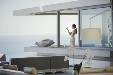 Frau mit Digitalkamera beim Fotografieren des Meerblicks auf dem Balkon eines modernen, luxuriösen Hauses - HOXF01267