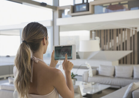Frau mit digitalem Tablet, die ein digitales Sicherheitssystem in einem modernen, luxuriösen Musterhaus im Wohnzimmer einstellt - HOXF01268