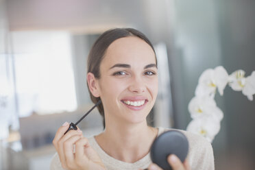 Porträt einer lächelnden, selbstbewussten Frau, die Wimperntusche mit einem Wimperntuschestift und einem Kompaktspiegel aufträgt - HOXF01276