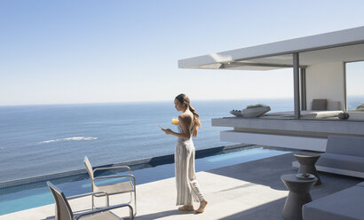 Frau, die mit ihrem Smartphone auf einer sonnigen, modernen, luxuriösen Terrasse mit Meerblick spazieren geht und SMS schreibt - HOXF01286